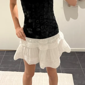 Vit volangkjol med spetsdetaljer - Supersöt vit kortkjol med volanger och fina spetsdetaljer. Kjolen har en luftig och lätt känsla, perfekt för varma dagar. Den har en feminin och lekfull look med flera lager och dekorativa sömmar.