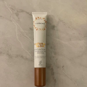 Lumene Natural Glow Primer - Lumene Natural Glow Moisturizing Illuminating Primer i en vit tub med bronsfärgat lock. Ger lyster och återfuktar huden, perfekt för normal till torr hud. Vegansk formula som boostar glow och gör basen jämnare. Tuben är smidig att använda och lätt att ta med.
