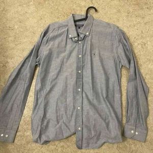 Grå skjorta från Tommy Hilfiger - Snygg grå skjorta från Tommy Hilfiger med klassisk button-down krage och diskret logga på bröstet. Skjortan har lång ärm, vita knappar och är tillverkad i mjuk bomull. Perfekt för dig som gillar stilrena och enkla plagg.