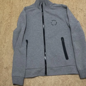 Grå ziptröja från BOSS med logga - Snygg grå ziptröja från BOSS med svart logga i cirkel på bröstet. Tröjan har hel dragkedja framtill, två fickor med dragkedja och ribbade muddar. Tillverkad i mjuk bomullsblandning, perfekt för lager-på-lager och chill vardagsstil.