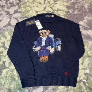 Mörkblå stickad tröja Polo Bear - Mörkblå stickad tröja från Polo Ralph Lauren med ikoniska Polo Bear-motivet framtill. Tröjan har rund hals, ribbade muddar och RL-broderi vid nederkanten. Tillverkad i mjuk bomull, perfekt för dig som vill ha en cool och klassisk look. Storlek S