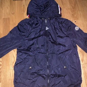 Mörkblå vindjacka från Moncler - Snygg mörkblå vindjacka från Moncler med huva och dragkedja framtill. Jackan har långa ärmar, snörning vid huvan och två fickor med tryckknappar. Moncler-logga på ärmen och coola rödvita detaljer på huvan. Perfekt för dig som gillar stilren och sportig look.