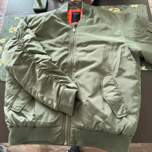 Olivgrön bomberjacka från Weekday - Snygg olivgrön bomberjacka från Weekday med klassisk orange insida. Jackan har ribbade muddar, dragkedja framtill och fickor med lock. Perfekt för dig som gillar streetstyle och vill ha en chill look.