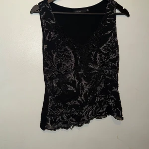 Svart blommig linnetopp från Vero Moda - Svart linnetopp från Vero Moda med blommigt mönster i grått och spetsdetaljer vid v-ringningen. Toppen är ärmlös och har en lätt vågig kant nedtill. Perfekt för dig som gillar en stilren men ändå detaljerad look.