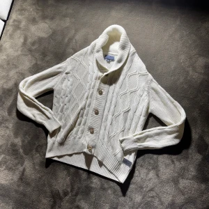 Vit vintage Cardigan❄️⛄️ - Säljer en krämvit stickad cardigan med grovt kabelstickat mönster, stora ribbade muddar och snygg sjalkrage. Koftan har träfärgade knappar framtill och en patch nertill. Perfekt för lager-på-lager under kyliga dagar. Storlek L men sitter som S/M