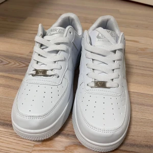 Nike Air Force 1 vita sneakers - Fräscha Nike Air Force 1 sneakers i helvitt skinn med klassisk låg siluett. Snygga detaljer som perforerad tå, snörning och metallbricka med 'AF1' på snörena. Ikonisk Nike-logga broderad på hälen och sidorna. Perfekt för dig som gillar clean och stilren look.