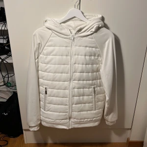 Vit quiltad pufferjacka med huva - Snygg vit pufferjacka med huva och quiltad design. Jackan har två sidofickor med dragkedja och hel dragkedja framtill. Materialet är mjukt och syntetiskt, perfekt för kyliga dagar. Stilren look med raka linjer och långärmade ärmar.