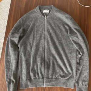 Grå kashmirkofta från Soft Goat - Grå kofta från Soft Goat i 100% kashmir med hel dragkedja framtill. Modellen har ribbade muddar och nederkant, samt långärmad passform. Perfekt för dig som gillar stilrena och mjuka plagg. Snygg att matcha med jeans eller chinos. Har ett lite håll på höger armen. Se bild