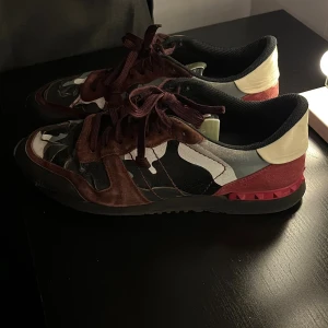 Valentino Rockrunner sneakers camouflage - Säljer ett par Valentino Rockrunner sneakers med grymt camouflagemönster i svart, grått, vitt och vinrött. Skorna har detaljer i mocka och skinn, röda nitar på hälen och platt sula. Snygga vinröda snören och rund tå. Perfekt för dig som vill sticka ut med lyxiga sneakers.