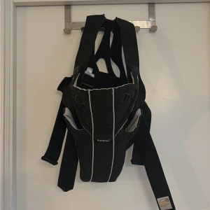 Svart BabyBjörn bärsele - Säljer en svart bärsele från BabyBjörn med vita kontrastsömmar. Selen har justerbara remmar och ergonomisk design för bekväm användning. Materialet är mjukt och slitstarkt, perfekt för att bära små barn nära kroppen. Enkel att ta på och av. Små fläckar som jag inte testat att få bort, i använt skick.