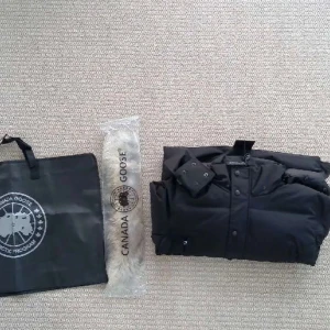 Svart dunjacka Canada Goose med päls - Säljer en svart dunjacka från Canada Goose med avtagbar äkta päls i beige och brun på huvan. Jackan har dragkedja och knappar framtill, samt flera praktiska fickor. Medföljer originalpåse och pälsdetalj. Perfekt för kalla vinterdagar.