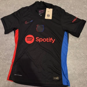 Svart FC Barcelona fotbollströja Nike - Säljer en svart FC Barcelona fotbollströja från Nike med röda och blå sidopaneler, Spotify-logga på bröstet och UNHCR-tryck på ryggen. Kortärmad modell i lätt, ventilerande Dri-FIT-material. Snygga detaljer med klubbmärke och Nike Swoosh på framsidan.