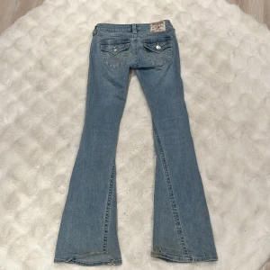 True Religion Joey Low Rise Flare Jeans - Snygga ljusblå jeans från True Religion, modell Joey Low Rise Flare. De har låg midja, utsvängda ben och klassiska bakfickor med lock och silverfärgade knappar. Jeansen är i denim med coola slitningar nertill och tydliga kontrastsömmar.
