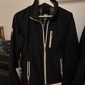 Marinblå vindjacka Helly Hansen XS - Svart vindjacka från Helly Hansen i storlek XS med vita dragkedjor och röda detaljer. Jackan har hög krage med broderad logga, meshfoder och flera fickor med dragkedja. Perfekt för dig som gillar sportig och clean stil.