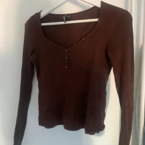 Brun ribbad långärmad topp Pieces - Brun långärmad topp från Pieces i ribbad bomullskvalitet. Toppen har v-ringning med dekorativa knappar framtill och sitter snyggt med en normal passform. Perfekt att styla med jeans eller kjol för en chill look.