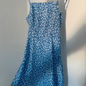 Blå blommig omlottklänning med knyt - Aldrig använd. Säljer en blå klänning med vita blommor, omlott och knyt i ryggen samt öppen rygg. Klänningen har smala axelband och är tillverkad i ett lätt material som ger en flowy känsla. Perfekt för sommarens alla tillfällen och har en feminin siluett.