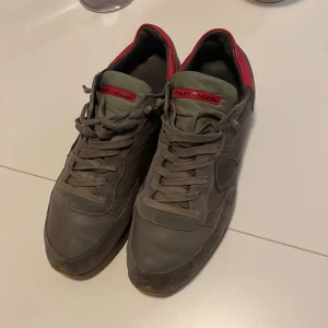 Grön Philippe Model sneakers med röd detalj - Snygga grå sneakers från Philippe Model med röd detalj på hälen och röd logga på plösen. Skorna har snörning, rund tå och platt sula. Materialmix av skinn och mocka ger en cool och urban vibe. Perfekta för dig som gillar stilrena och unika sneakers.