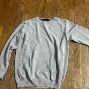 Ljusgrå stickad tröja Tommy Hilfiger - Snygg ljusgrå stickad tröja från Tommy Hilfiger med diskret logga på bröstet. Rund hals och ribbade muddar vid ärmslut och nederkant. Perfekt för dig som gillar klassisk stil med en modern touch.