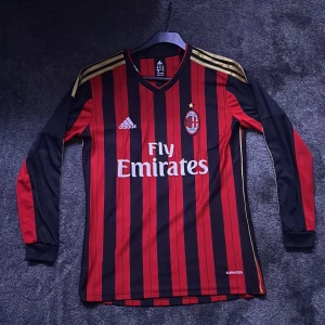 AC Milan långärmad fotbollströja Adidas - Säljer en officiell AC Milan långärmad matchtröja från Adidas med klassiska röda och svarta ränder, guldiga axelstreck och broderat klubbmärke med stjärna. Tröjan har Fly Emirates-tryck på bröstet och är tillverkad i lätt climacool-material. Min pappa köpte den i Milan shoppen i Milano. 