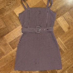 Rutig rosa klänning från Pull&Bear - Supersöt kortklänning från Pull&Bear i rosa och svart rutigt mönster. Klänningen har axelband, markerad midja med matchande bälte och en rak, figurnära passform. Perfekt för dig som gillar en trendig och lekfull stil.