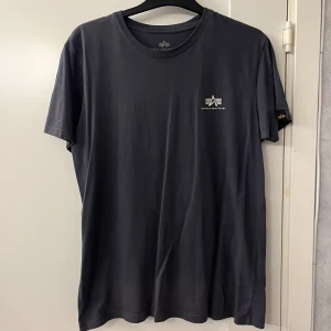 Mörkgrå t-shirt Alpha Industries L - Snygg mörkgrå t-shirt från Alpha Industries i storlek L. Klassisk rund hals och korta ärmar, med diskret logga tryckt på bröstet och en liten patch på ärmen. Tillverkad i mjuk bomull som känns skön mot huden. Fint skick på den, snygg t-shirt som passar till nästan allt. Hör av er om ni har frågor!