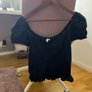 Svart blus med puffärm och volang - Säljer en mörkblå blus från Nelly med puffiga korta ärmar och smockad överdel. Blusen har en söt volangkant nedtill och en lätt rynkad struktur som ger en cool och avslappnad vibe. Perfekt för dig som gillar trendiga detaljer och vill sticka ut lite extra.