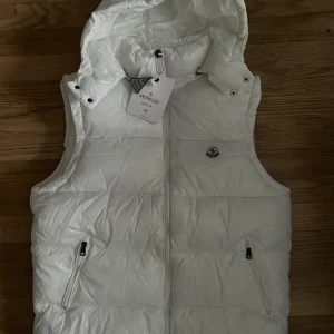 Vit dunväst från Moncler med huva - Säljer en snygg vit dunväst från Moncler i storlek M. Västen har en quiltad design, praktisk huva och två fickor med dragkedja. Moncler-logga på bröstet och dragkedja framtill. Perfekt för lager-på-lager och stilrena outfits.