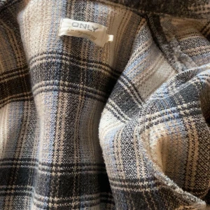 Rutig jacka från ONLY - Säljer en superfin rutig jacka från ONLY. Den har en klassisk krage och knappar framtill. Perfekt för höst och vår med sitt mysiga rutmönster i svart, grått och beige. Jackan är långärmad och har en normal passform. Perfekt för att ge din outfit en stilren touch!