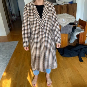 Rutig kappa med bälte - Säljer en superfin rutig kappa i beige, brun och svart. Den har ett bälte i midjan för en snygg passform och långa ärmar. Perfekt för höst och vår när det är lite kyligt ute. Kappan är i bra skick och redo för nya äventyr!