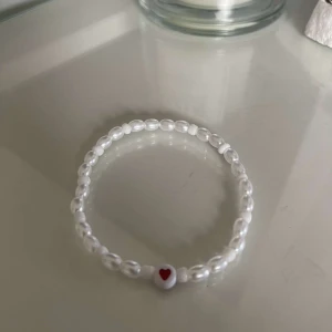 Pärlarmband med hjärta - Säljer ett supergulligt armband med vita pärlor och en liten röd hjärtpärla i mitten. Perfekt för att ge en söt touch till din outfit! Passar till alla tillfällen och är one size.