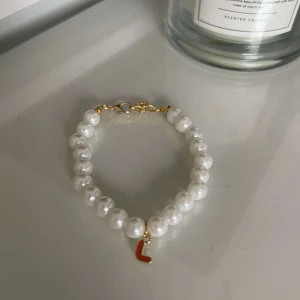 Pärlarmband med bokstavsberlock - Snyggt pärlarmband med vita pärlor och en liten guldberlock formad som bokstaven 'L'. Perfekt för att ge en personlig touch till din outfit. Armbandet har en enkel guldfärgad låsning.