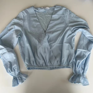 Ljusblå omlottopp - Säljer en superfin ljusblå omlottopp med långa ärmar. Toppen har en v-ringad design och elastiska muddar vid ärmslut och nederkant, vilket ger en snygg passform. Perfekt för både vardag och fest!