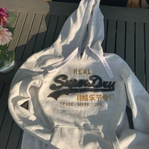 Ljusgrå Superdry Hoodie med glittrigt tryck - Mycket skön och i fint skick förutom en liten fläck på nedre delen av tröjan och en liten lös tråd i trycket som fanns där från början.