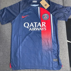Blå PSG fotbollströja - Säljer en snygg blå PSG fotbollströja från Nike. Den har en röd och vit rand på framsidan och är kortärmad. Perfekt för alla fotbollsfans som vill visa sitt stöd för Paris Saint-Germain. Tröjan är i nyskick och har Qatar Airways som sponsorlogga på bröstet.