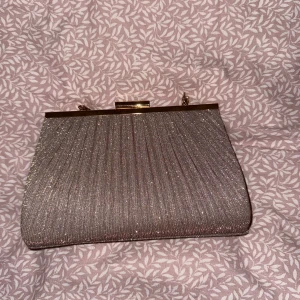 Glittrig kuvertväska med gulddetaljer - Elegant glittrig kuvertväska med gulddetaljer och en stilren design. Perfekt att ha till tex balen eller andra festliga tillfällen. Väskan har en praktisk öppning och en kedja för att bäras över axeln. Använd en gång