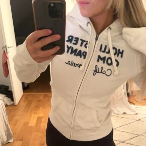 Vit hoodie från Hollister - Säljer en vit hoodie från Hollister med dragkedja och blå text på bröstet. Den har en justerbar huva med snören och långa ärmar. Perfekt för en avslappnad stil. Storlek L men passar s