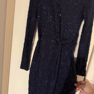 Mörkblå glittrig festklänning - Elegant mörkblå klänning med glittriga detaljer. Den har långa ärmar och en knytning i midjan som ger en smickrande passform. Lång och delar sig framifrån. Perfekt för festliga tillfällen. Endast använt vid två tillfällen.