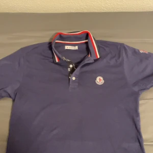 Blå pikétröja från Moncler - Snygg blå pikétröja från Moncler med klassisk krage och röd-vit randig detalj. Tröjan har en broderad Moncler-logga på bröstet och knappar framtill. Perfekt för en stilren look.