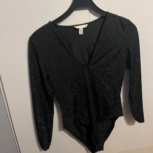 Svart glittrig bodysuit - Snygg svart bodysuit med glittrigt material och v-ringad design. Perfekt för en kväll ute eller fest. Långärmad och figurnära passform. Sjukt snygg på!!