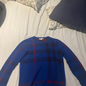 Burberry tröja i kashmire - Säljer en stilren blå tröja från Burberry med ett klassiskt rutmönster i rött och svart. Tröjan är långärmad och tillverkad i Kashmir och bomull, vilket gör den både bekväm och elegant. Perfekt för att ge en touch av lyx till din garderob.