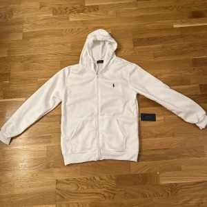 Vit hoodie från Polo Ralph Lauren - Säljer en stilren vit hoodie från Polo Ralph Lauren med dragkedja och det klassiska logotypbroderiet på bröstet. Perfekt för en avslappnad look.