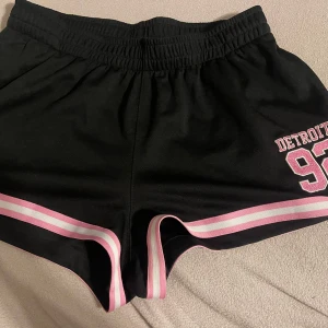 Svarta shorts med rosa detaljer från FB Sister/ new yorker - Snygga svarta shorts från FB Sister med rosa och vita ränder längs kanterna. De har elastisk midja och texten 'Detroit 92' tryckt på ena benet. Perfekta för en avslappnad stil.