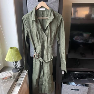 Olivgrön klänning från MNG - Snygg olivgrön klänning från MNG med knappar framtill och ett bälte i midjan. Klänningen har långa ärmar och en klassisk krage, perfekt för en stilren look.