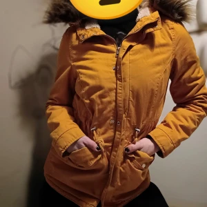 Gul parkajacka med pälskrage - Snygg gul parkajacka med fuskpäls luva och dragkedja. Jackan har snören man kan justera för att framhäva midjan och praktiska fickor framtill. Perfekt för kyligare dagar.
