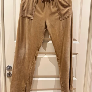 Beige mjukisbyxor från Juicy Couture - Säljer ett par snygga beige mjukisbyxor från Juicy Couture. Byxorna har en broderad logga på framsidan och praktiska fickor både fram och bak. De är tillverkade i ett mjukt material och har en bekväm passform med snörning i midjan.