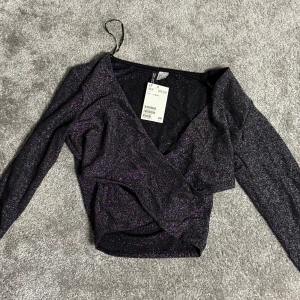 Svart glittrig omlottopp från H&M - Snygg svart glittrig omlottopp från H&M med långa ärmar. Perfekt för en festlig kväll. Toppen har en elegant omlottdesign som ger en smickrande passform.