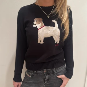 Svart tröja med hundmotiv från Ralph Lauren - Säljer en svart tröja från Ralph Lauren med ett stickat hundmotiv i beige och brunt framtill. Tröjan har långa ärmar och en rund halsringning. 