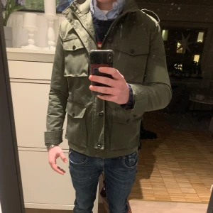 Grön fieldjacket - Säljer en stilren grön field jacket med praktiska fickor och huva. Jackan har långa ärmar och knappar framtill. Perfekt för kyligare dagar. Jackan kan användas året om och är i nyskick.