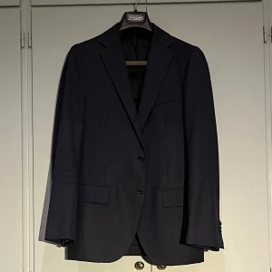 Mörkblå kostym från Gaiola - Mörkblå kostym med diskreta ränder från Gaiola. Kavajen har klassiska slag och knappar framtill. Skick 7/10. Storlek 46
