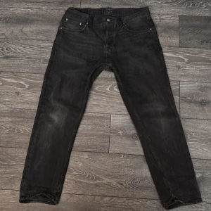 Svarta jeans från Nudie Jeans - Snygga svarta jeans från Nudie Jeans med klassisk femficksdesign och knappgylf. Perfekta för en stilren look. Märkesdetaljer på knappar och etikett.